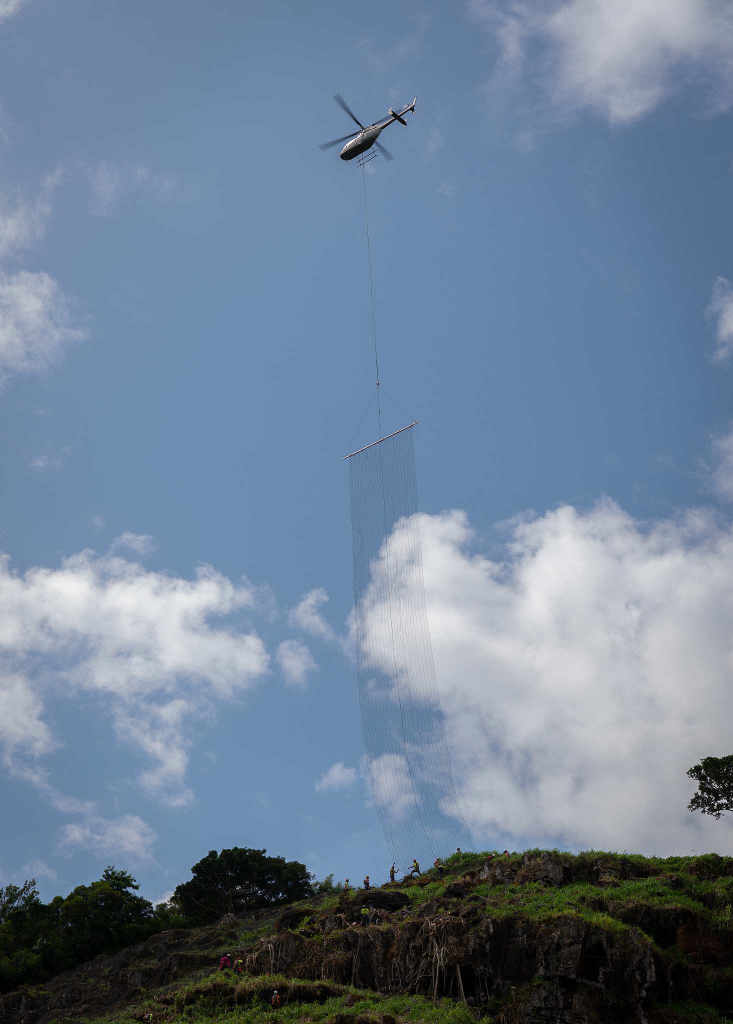 A helicopter lowers a 24 by 150 foot length of  rockfall netting next to workers visible on the ridgeline above Waimea Bay May 17, 2026. The 150-foot-long length of netting weighs about 2000 pounds. (Craig Fujii/Civil Beat/2026)